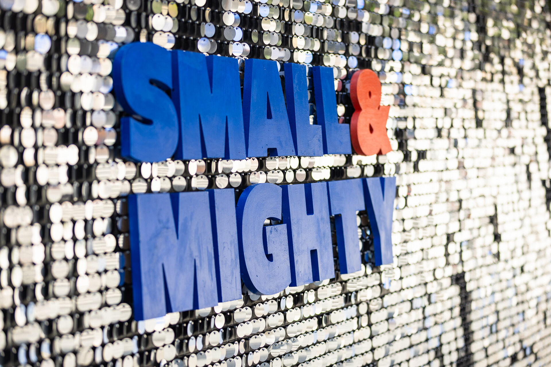 &ldquo;Small & Mighty&rdquo; sign in blue and orange lettering on a shimmering silver sequin wall